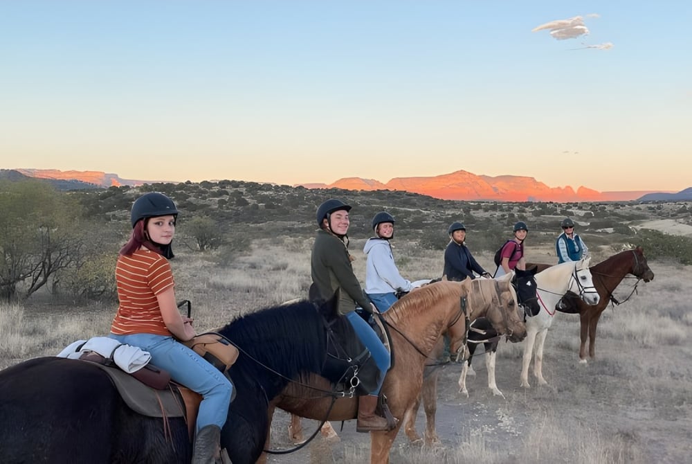 Schüler der Verde Valley School reiten auf Pferden durch eine landschaftlich reizvolle Wüstenlandschaft mit Bergen im Hintergrund.
