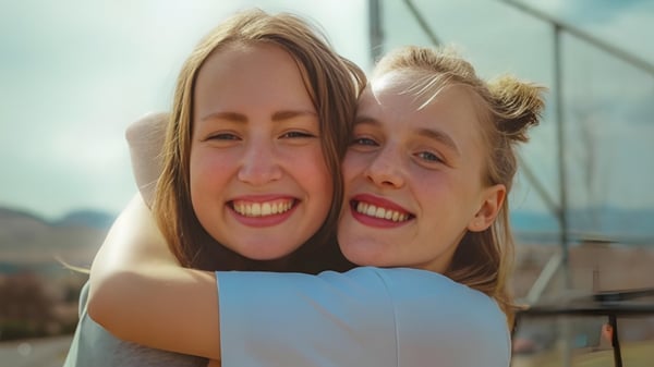 Zwei Schülerinnen mit blonden Haaren umarmen sich lächelnd vor einer landschaftlichen Kulisse auf dem Campus der Vernon Secondary School.