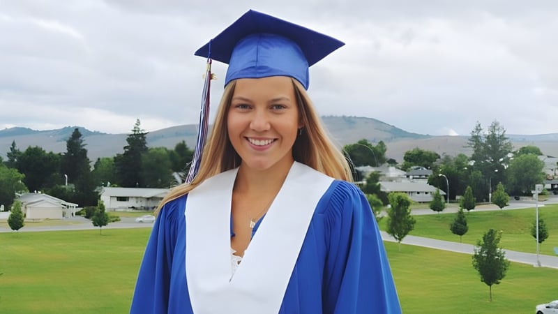 Eine junge Frau im blauen Abschlussgewand steht vor Bergen auf dem Gelände der Vernon Secondary School.