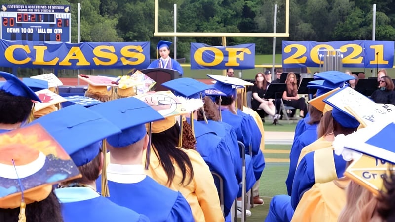 Eine Gruppe von Absolventinnen und Absolventen der Vernon Township High School steht in blauen und goldenen Talaren vor einem Banner mit der Aufschrift Class of 2021 auf einem Sportplatz.