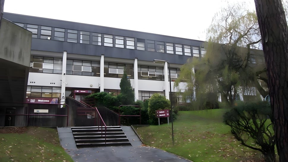 Das mehrstöckige graue Gebäude auf dem Campus des Victoria College Belfast ist von Bäumen und Grün umgeben.