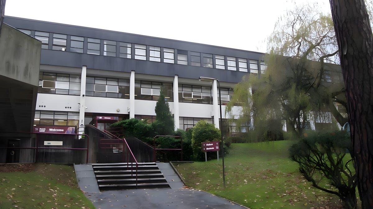 Das mehrstöckige graue Gebäude auf dem Campus des Victoria College Belfast ist von Bäumen und Grün umgeben.