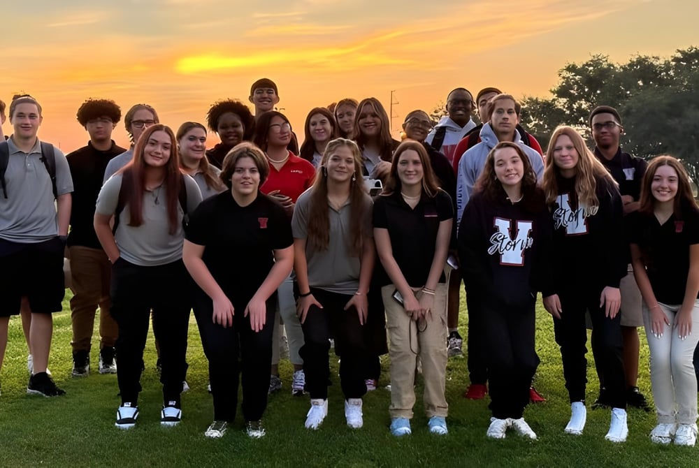 Eine Gruppe von Schülerinnen und Schülern der Victory Christian Academy steht gemeinsam im Freien vor einem Sonnenuntergang mit Bäumen im Hintergrund.