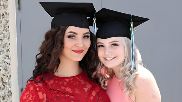 Zwei Schülerinnen der W.F. Herman Academy Secondary School posieren lächelnd mit Abschlusskappen vor einer grauen Wand.