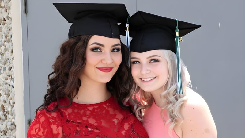 Zwei Schülerinnen der W.F. Herman Academy Secondary School posieren lächelnd mit Abschlusskappen vor einer grauen Wand.