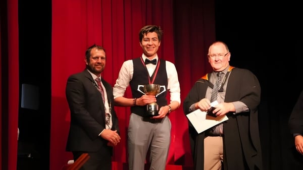 Drei Männer stehen auf der Bühne mit rotem Vorhang, einer hält einen Pokal bei einer Preisverleihung am Waihi College.