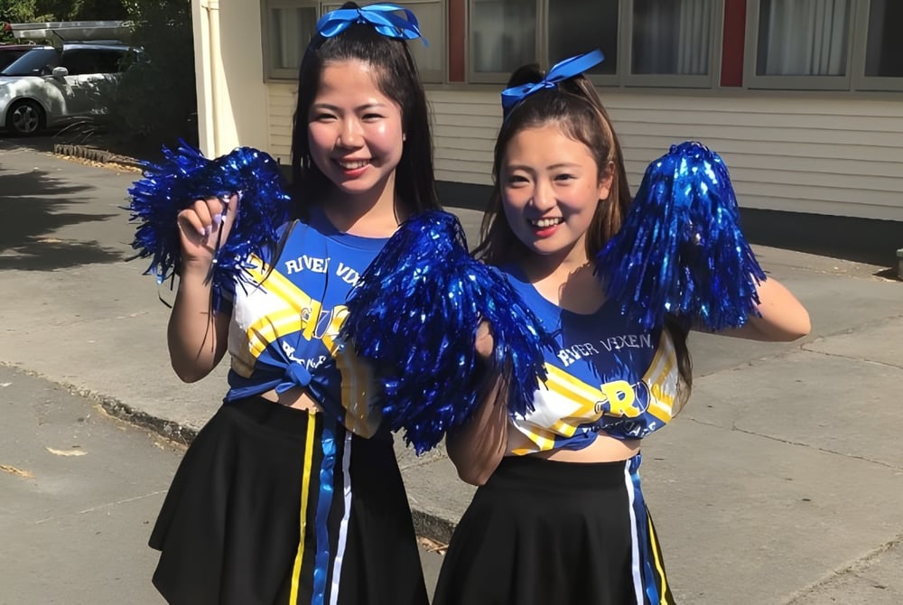 Zwei Schülerinnen in Cheerleader-Uniformen stehen mit Pompons vor einem Gebäude auf dem Campus des Waimea College.