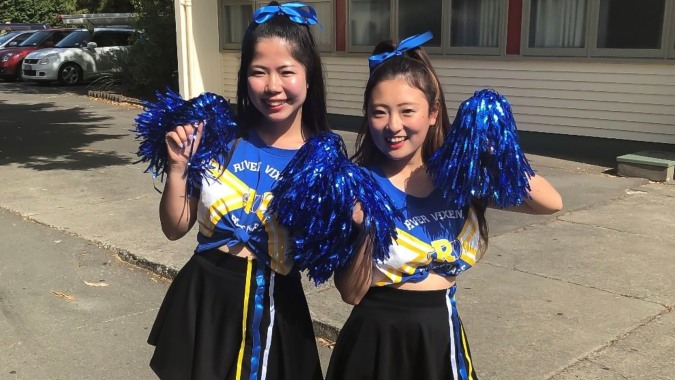 Zwei Cheerleader der Waimea College stehen in blauen und gelben Uniformen mit Pompons vor einem Gebäude.