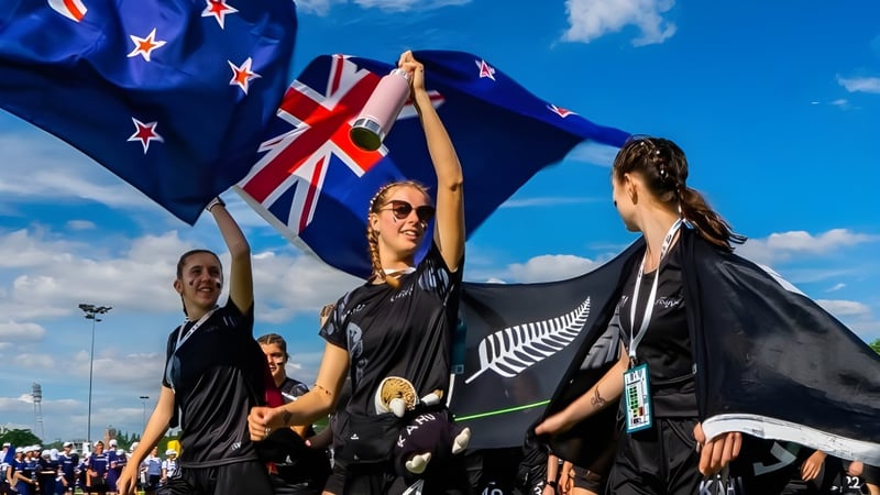 Eine Gruppe von Schülerinnen und Schülern des Waitakere College winkt mit neuseeländischer Flagge vor blauem Himmel mit Union Jack und Silberfarnflagge.