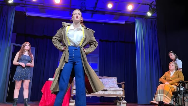 Eine Frau im langen beigen Mantel steht auf der Bühne des Waiuku College, umgeben von anderen Frauen in bunten Kleidern.