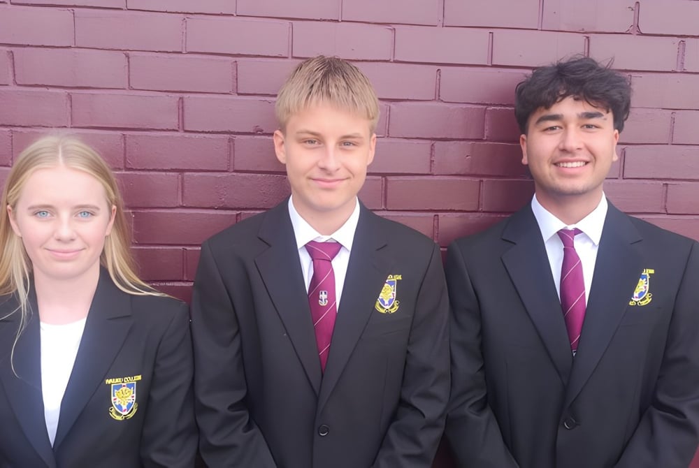 Drei Schülerinnen und Schüler in Schuluniform stehen vor einer roten Backsteinmauer am Waiuku College.