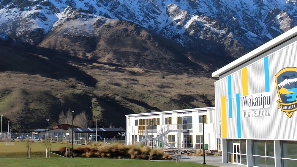 Das bunte Gebäude der Wakatipu High School steht vor schneebedeckten Bergen und grüner Landschaft.