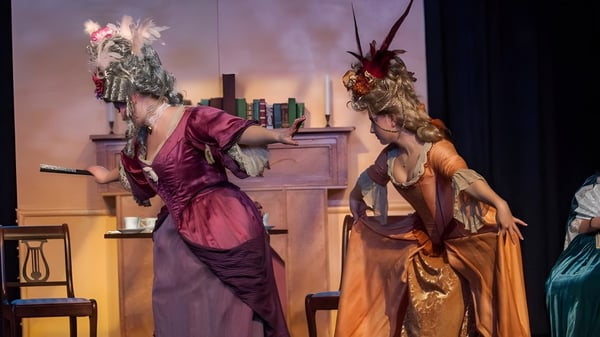 Zwei farbenfroh gekleidete Performer stehen auf der Bühne des Walkerville Collegiate Institute vor einer Kulisse mit Regalen und Möbeln.