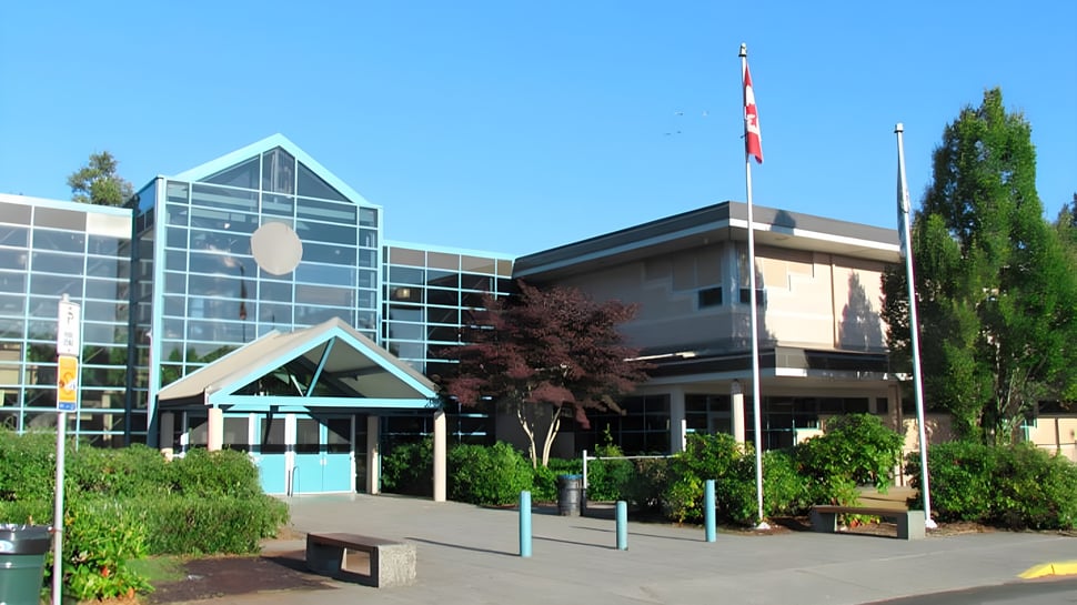 Das moderne Glas- und Betongebäude der Walnut Grove Secondary School mit kanadischer Flagge im Vordergrund und umgeben von Bäumen ist zu sehen.