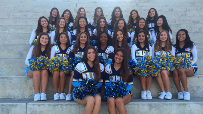 Eine Gruppe junger Schülerinnen in Cheerleading-Uniformen mit Pompons posiert vor einer Betonwand auf dem Gelände des Walnut Valley School District.