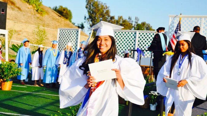 Absolventen des Walnut Valley School District stehen in weißen Kleidern und Hüten auf einer Wiese vor Gebäuden und Bäumen.