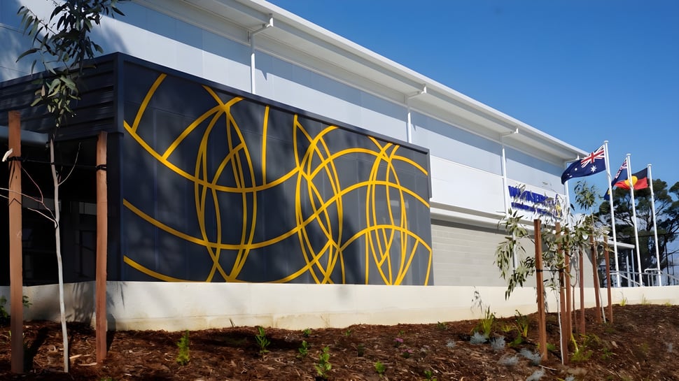 Das große weiße Gebäude mit einem gelben abstrakten Wandbild auf dem Campus des Wanneroo Secondary College vor blauem Himmel.