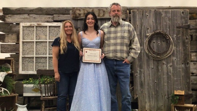 Drei Personen stehen vor einer rustikalen Holzwand auf dem Gelände der Warner School.