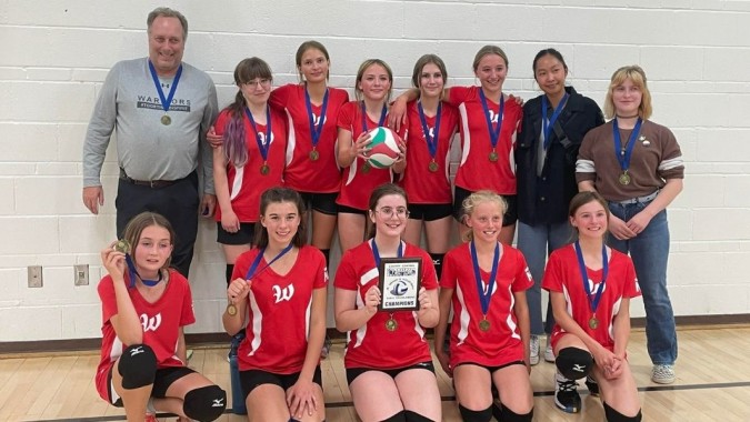 Eine Gruppe junger Volleyballspielerinnen in roten Trikots posiert mit ihren Trainern und einem Pokal in der Sporthalle der Warner School.