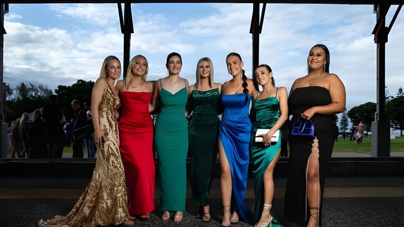 Sechs Schülerinnen der Warners Bay High School stehen in Abendkleidern vor einer landschaftlichen Kulisse mit bewölktem Himmel.