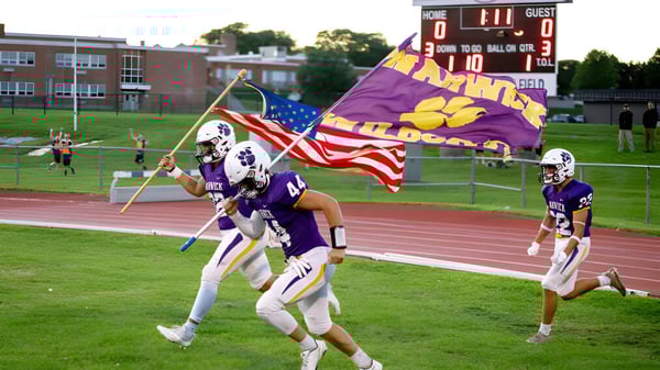 Eine Gruppe von Footballspielern der Warwick Valley High School trägt auf dem Spielfeld Fahnen und Banner ihres Teams und Landes.