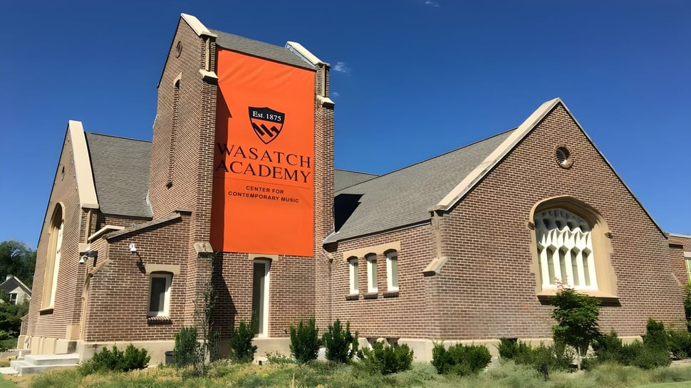 Das große Backsteingebäude mit einem orangefarbenen Banner der Wasatch Academy vor klarem blauem Himmel und grüner Umgebung.