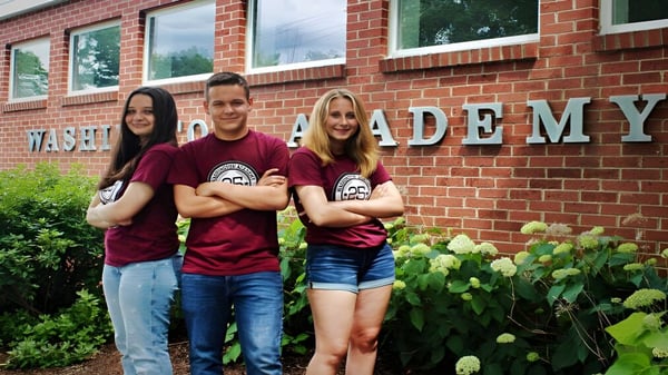 Drei Schülerinnen und Schüler stehen vor einem Backsteingebäude der Washington Academy und posieren für ein Foto.