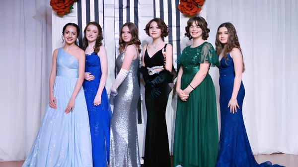 Eine Gruppe von sechs Schülerinnen in formellen Abendkleidern steht vor rotem Blumendekor auf dem Campus der Webb School.