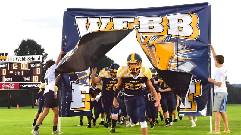 Eine Gruppe von Fußballspielern betritt das Spielfeld durch ein großes Banner mit dem Schriftzug Webb auf dem Campus der Webb School.