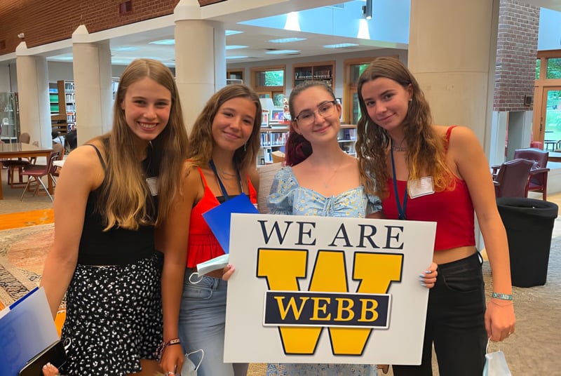 Vier Schülerinnen der Webb School halten gemeinsam ein Schild mit der Aufschrift ‚WE ARE WEBB‘ in einem Café.