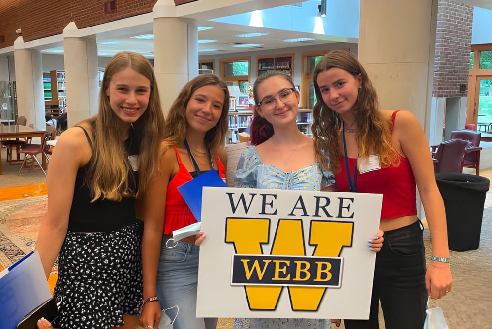 Vier Schülerinnen der Webb School halten gemeinsam ein Schild mit der Aufschrift ‚WE ARE WEBB‘ in einem Café.