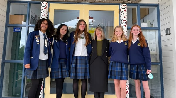 Eine Gruppe von sechs Schülerinnen in Schuluniformen steht vor einem Gebäude des Wellington East Girls’ College mit verzierten architektonischen Details.