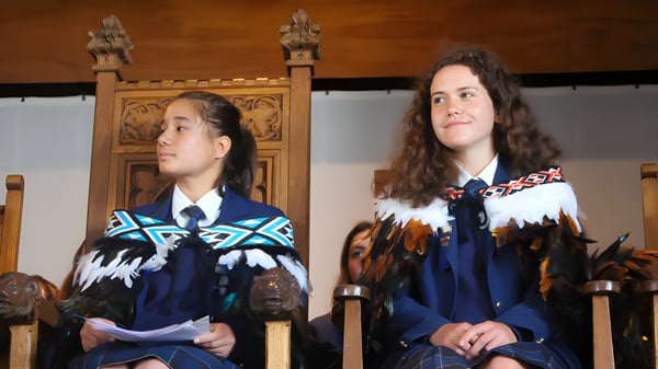 Zwei Schülerinnen des Wellington East Girls’ College tragen traditionelle Kleidung vor einer hölzernen Struktur mit Schnitzereien.