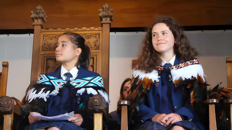 Zwei Schülerinnen des Wellington East Girls’ College tragen traditionelle Kleidung vor einer hölzernen Struktur mit Schnitzereien.
