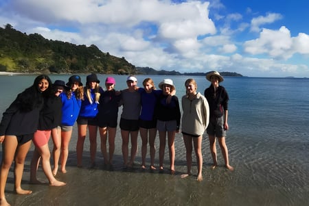 Eine Gruppe von Schülerinnen des Wellington Girls' College steht gemeinsam am Strand mit Bergen im Hintergrund.