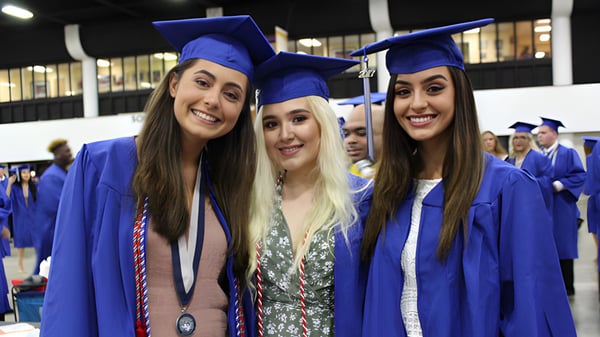 Drei Schülerinnen der Wellington Heights Secondary School feiern in blauen Abschlussroben und Mützen ihre Graduierung.