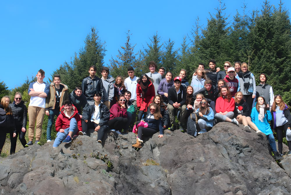 Eine Gruppe von Schülerinnen und Schülern der Wellington Secondary School steht auf einem Felsen umgeben von immergrünen Bäumen unter klarem blauem Himmel.