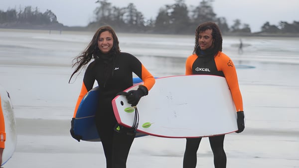 Zwei Personen in Neoprenanzügen mit Surfbrettern stehen am Strand vor einem bewaldeten Hintergrund auf dem Gelände der Wellington Secondary School.