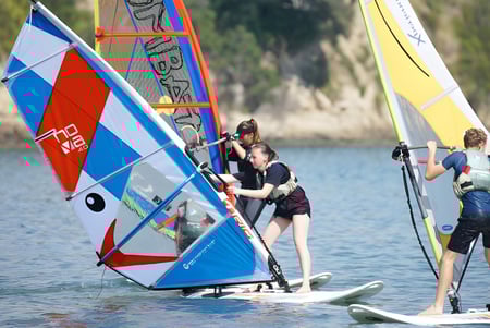 Segelboote und Windsurfer fahren auf einem ruhigen Gewässer in der Nähe des Wentworth College, umgeben von bewaldeter Landschaft.