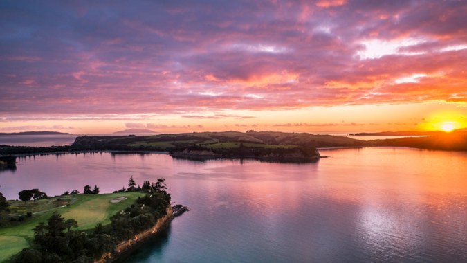 Ein ruhiger See mit grünen Hügeln und Bergen im Hintergrund bei einem bunten Sonnenuntergang am Wentworth College.