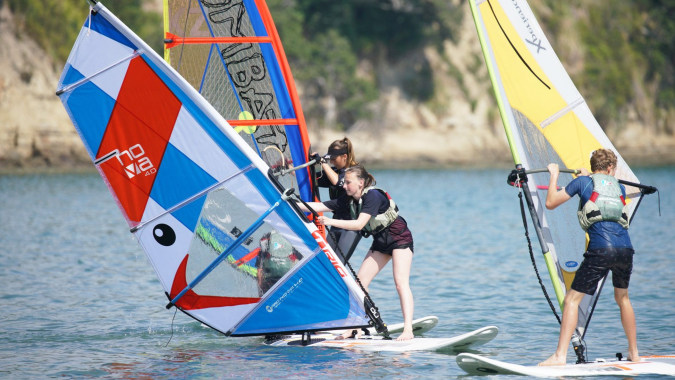 Schüler des Wentworth College segeln mit bunten Windsurfboards auf einem Gewässer vor bewaldeter Kulisse.