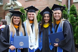 Vier Absolventinnen in Talar und Barett halten ihre Diplome vor einem Backsteingebäude auf dem Campus des Wesley College.