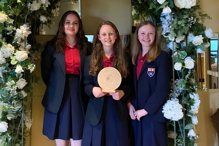 Drei Absolventinnen des Wesley College stehen gemeinsam vor einem floral geschmückten Bogen und halten ihr Diplom.