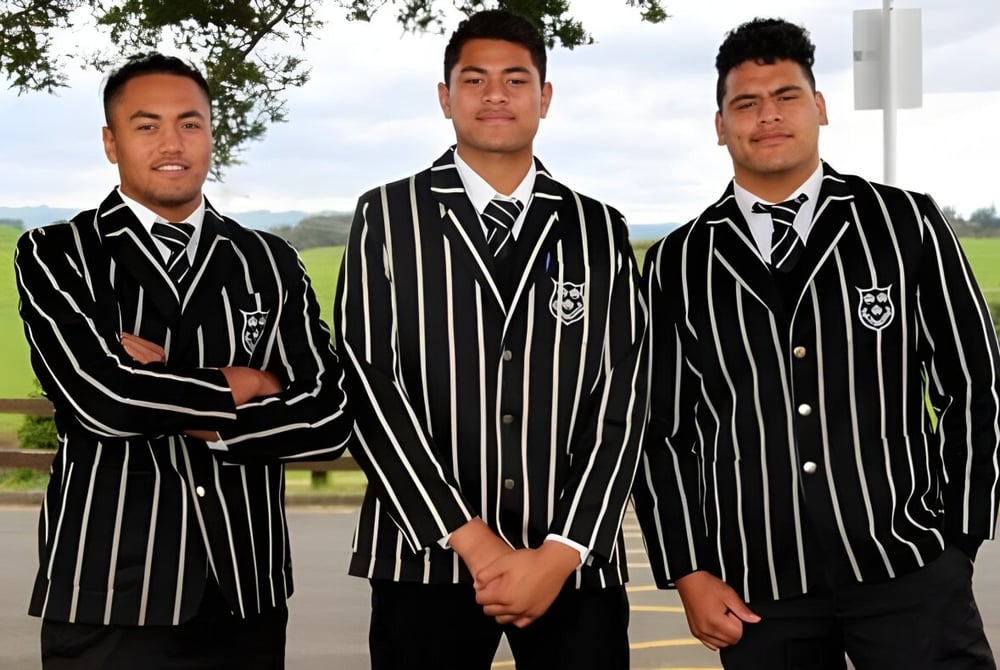 Drei Schüler des Wesley College stehen im Freien auf einer Wiese vor Bäumen in formellen schwarz-weißen Anzügen.