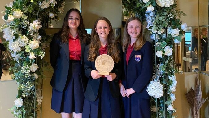 Drei Schülerinnen in Schuluniformen stehen im Blumenbogen und halten eine Auszeichnung am Wesley College.