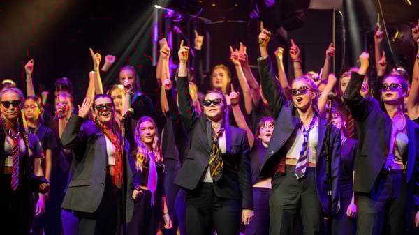 Eine große Gruppe formell gekleideter Schüler auf der Bühne der West Buckland School mit erhobenen Armen unter buntem Licht.