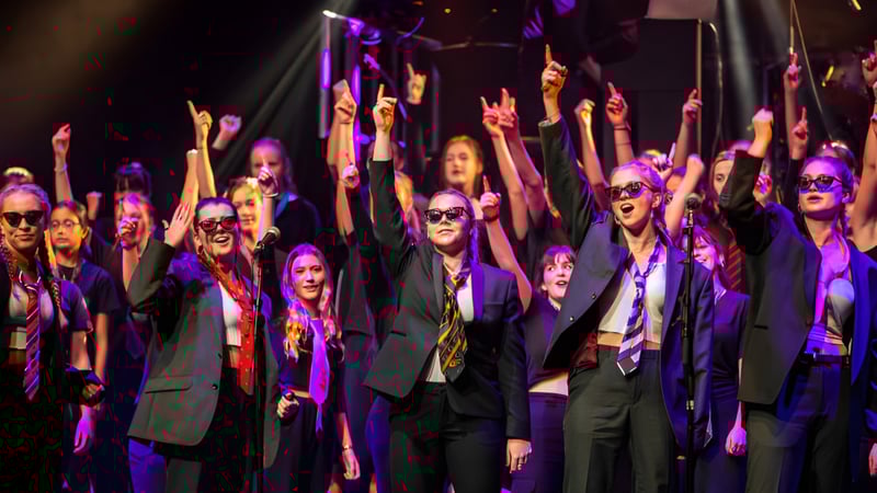 Eine große Gruppe formell gekleideter Schüler auf der Bühne der West Buckland School mit erhobenen Armen unter buntem Licht.