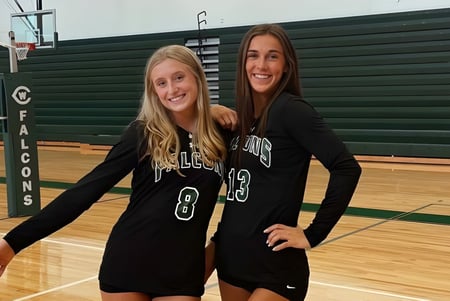 Zwei Schülerinnen der West Catholic High School stehen in Sporttrikots in einer Turnhalle vor einer grünen Wand und lächeln.