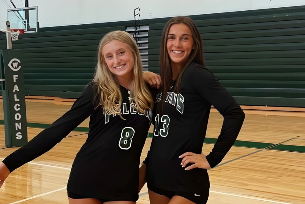 Zwei Schülerinnen der West Catholic High School stehen in Sporttrikots in einer Turnhalle vor einer grünen Wand und lächeln.