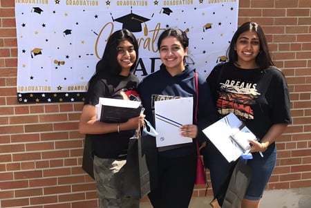 Drei Schülerinnen des West Humber Collegiate Institute stehen vor einer Abschlusskulisse und halten stolz ihre Diplome.